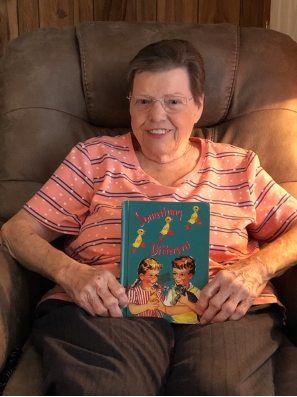 Jane holds up the 'Something Different' book that she purchased at a yard sale with the hope of one day learning how to read it. 
