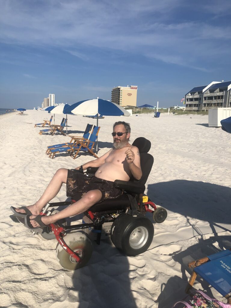 Tim sits on the beach in the beach wheelchair that Enhabit Cares was able to provide him with.