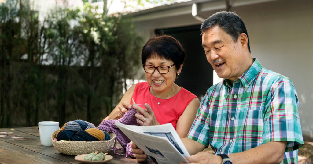 Image shows two older adults smiling and reading a newspaper together outside