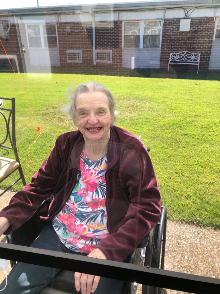 Granny smiles outside of her nursing facility in 2020 with a plastic screen separating her from having direct contact with her family.