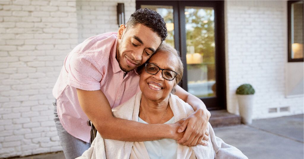 A caregiver huges his loved one from the side and they both smile.