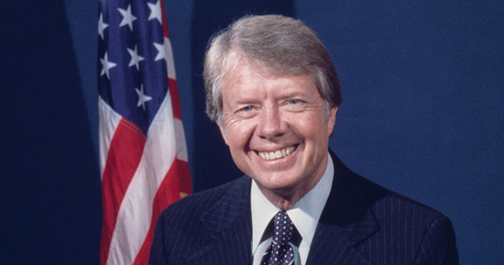 Former President Jimmy Carter smiling in front of an American flag. 