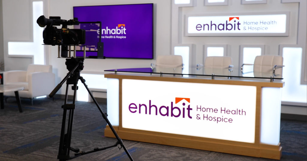 The Enhabit Enrichment Community (EEC) has a broadcast studio for live and on-demand trainings. This picture shows a film camera and three chairs sitting at a table with the Enhabit logo. 