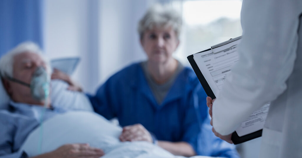 A physician holds a clipboard and stands in front of a male patient in a hospital bed with their oxygen mask and their wife, mother or daughter. This resembles a discussion with a patient and their loved one about hospice indicators and signs the patient could benefit from hospice care. 