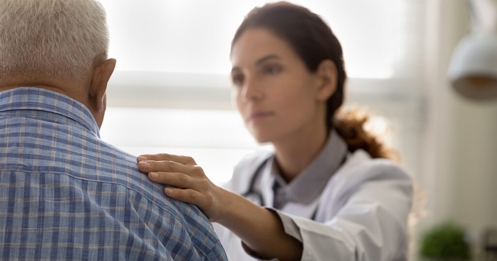A female doctor puts her hand on an older male's shoulder, demonstrating or representing how to talk to patients about hospice