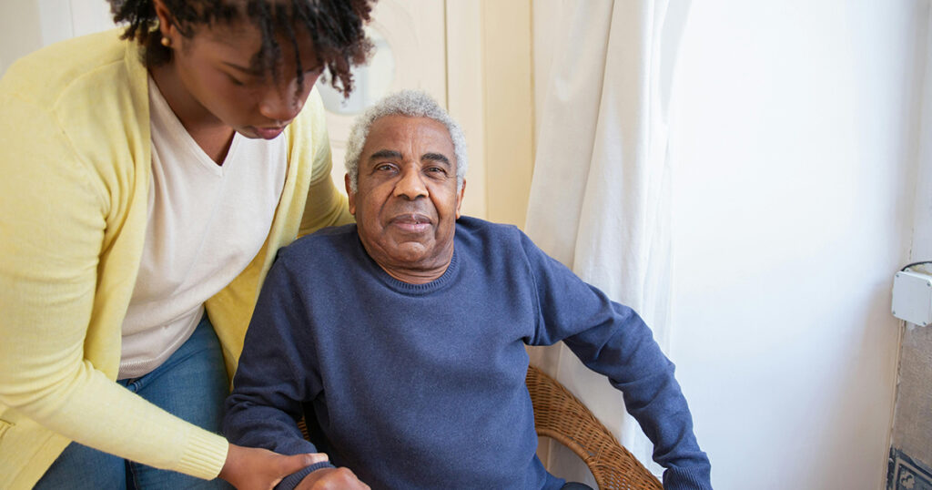 This image shows a woman in a yellow cardigan helping an older man in a blue sweater out of a chair. This highlights how caregivers need assistance and how home health and hospice services can benefit caregivers