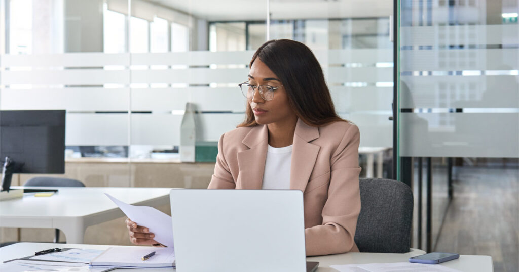 A woman wearing a peach-colored suit sits at an office desk, reading a document and working on her computer, as a branch director would do.