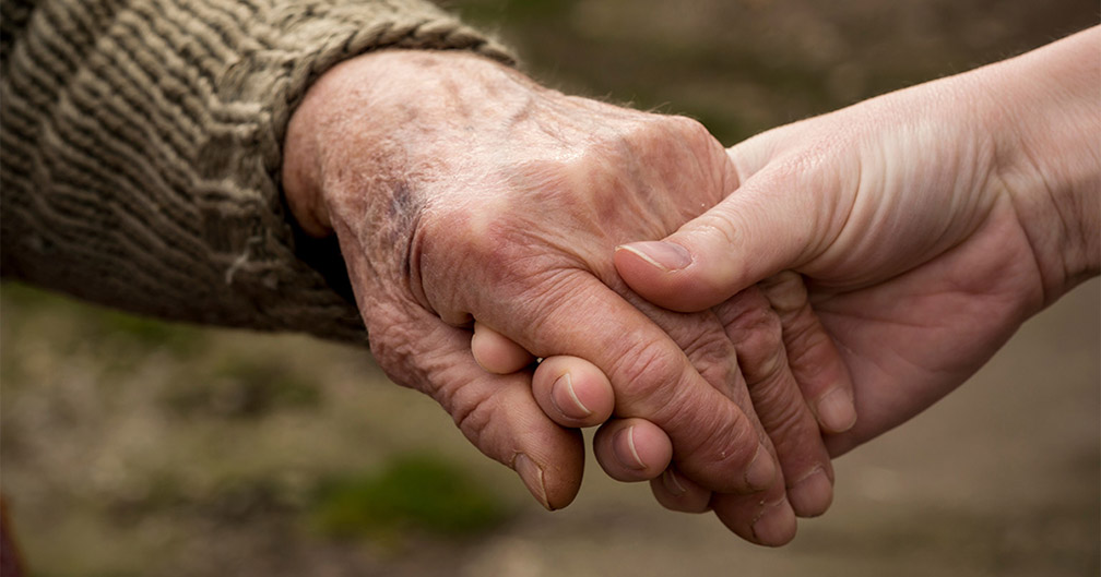 An older hand on the left holds a younger hand on the right, simulating support during a hospice conversation with your loved one